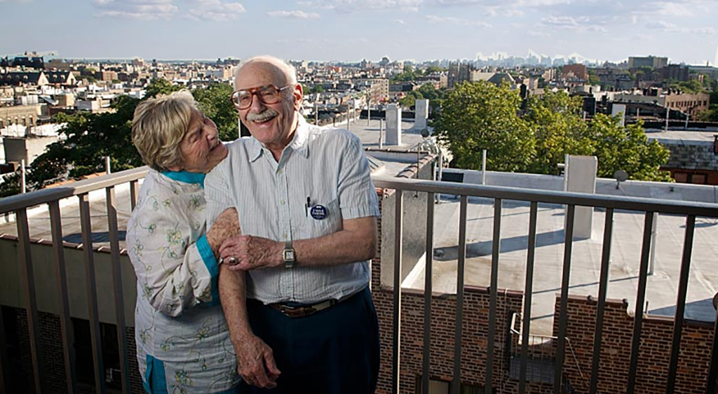 An older man wearing glasses cheerfully stands next to his wife on their balcony at The New Jewish Home Kittay apartments