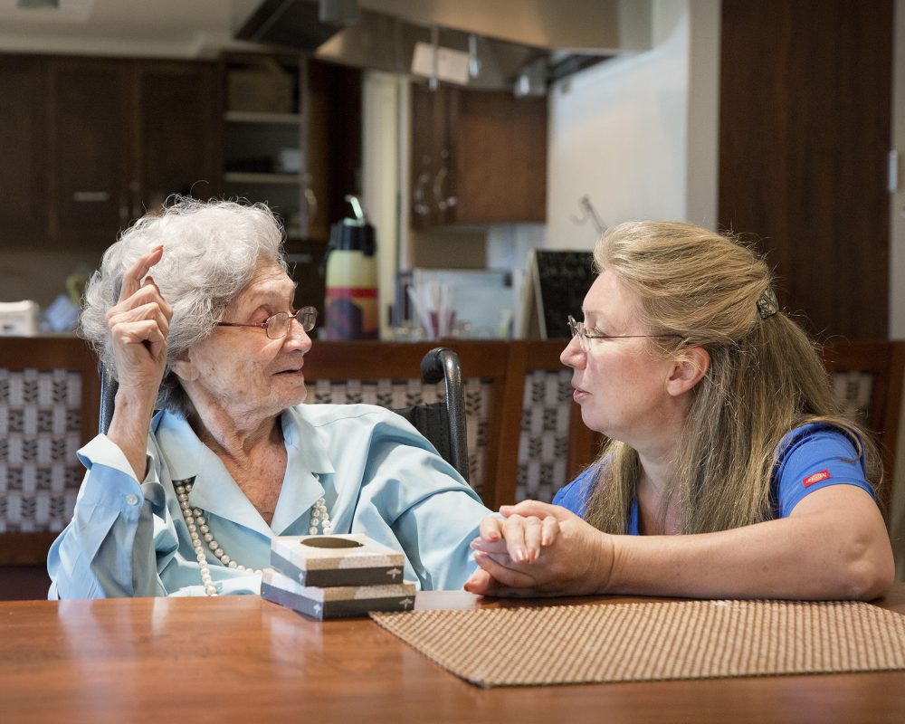 An elderly woman with gray hair and glasses sits at a table, smiling while holding the hand of a younger woman with glasses and long hair, who is wearing a blue shirt. They appear to be in a cozy, home-like setting, engaging in a warm conversation.