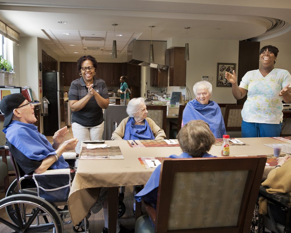 A group of elderly people sit at a dining table in a communal area, smiling and engaging with each other. Two caregivers stand nearby, clapping and smiling. The room is well-lit with a kitchen in the background, and the atmosphere appears joyful and lively.