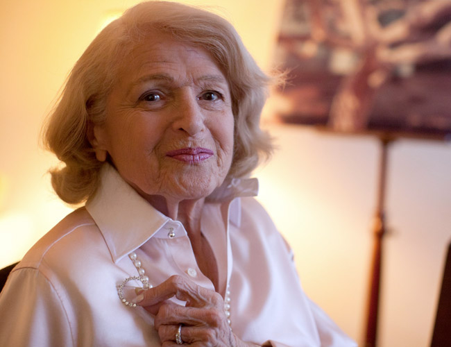 An elderly woman with light hair wearing a white blouse is smiling gently. She is seated indoors in a warmly lit room and holding a piece of jewelry on her blouse. The background is slightly out of focus, showing a framed picture and a lamp.