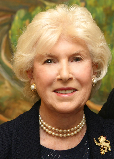 Portrait of an elderly woman with short, curly white hair, wearing pearl earrings, a pearl necklace, and a dark blouse. She has a friendly expression and is posed in front of a greenish, blurred background.