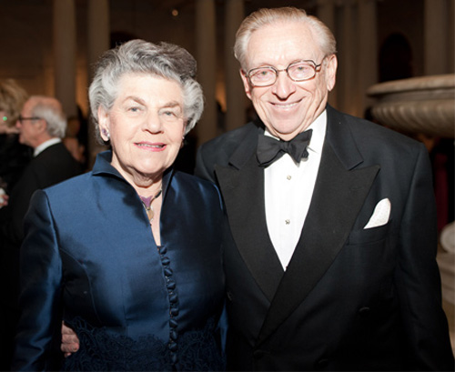 Older woman with grey hair wearing a blue dress stands next to an older man with graying hair and glasses wearing a black tux