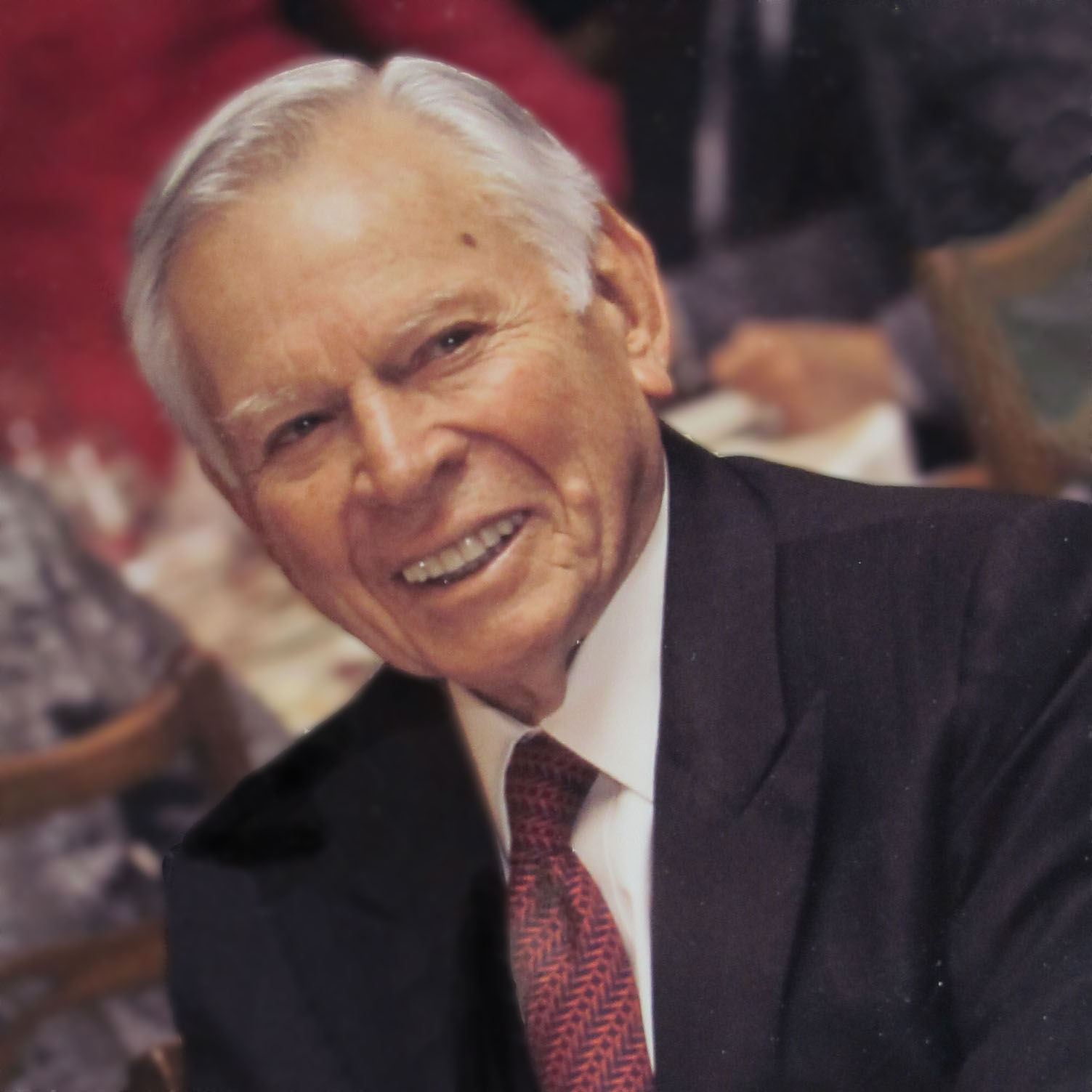 An elderly man with white hair wearing a black suit, white shirt, and red patterned tie smiles warmly at the camera. The background is softly blurred, suggesting an indoor setting, possibly an event or gathering.