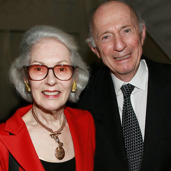 An elderly couple is smiling at the camera. The woman is wearing glasses, a red blazer, and a gold necklace with a large pendant. The man is wearing a black suit with a white shirt and a patterned tie. They appear to be indoors.