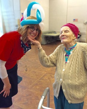 Volunteer at The New Jewish Home wearing a balloon hat and bends over so that an older lady using a walker can admire it
