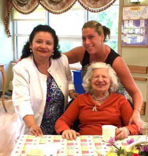 Two Female volunteers at The New Jewish Home talk to a woman sitting in a chair, drinking tea