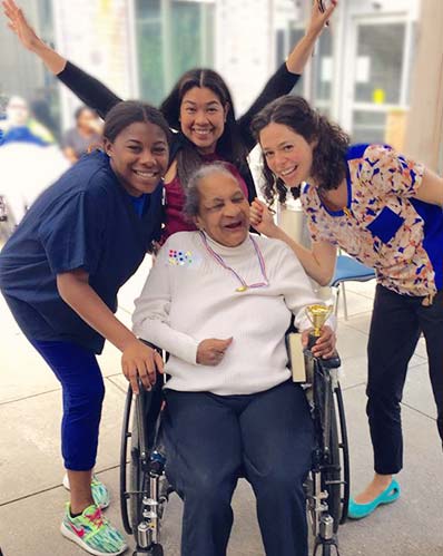 Three volunteers are standing and cheering with an elderly Hispanic woman in a wheelchair at the New Jewish Home