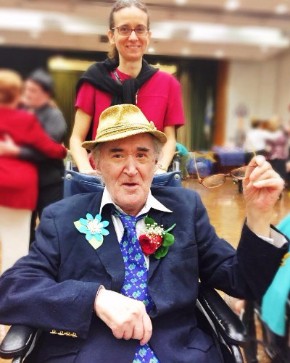 Volunteer at The New Jewish Home pushes a man in a wheelchair wearing a blue suit and tie as well as two bright flowers