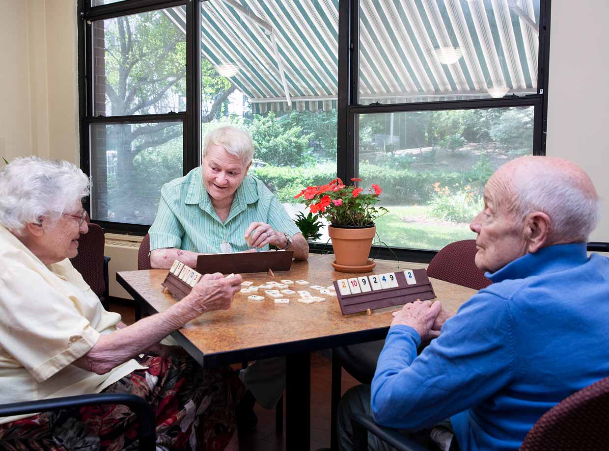 Three older adults sit at a table with a vase of red flowers by a window as they play dominoes at the New Jewish Home.