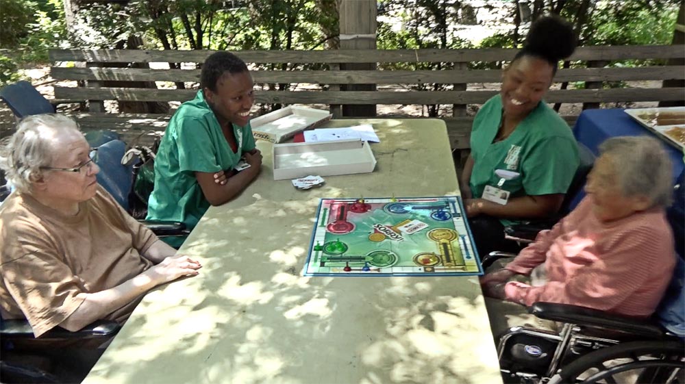 Four people are gathered around a table playing a board game on a sunny day. Two elderly individuals in wheelchairs sit opposite two caregivers wearing green uniforms. They appear to be enjoying themselves, smiling and engaging in conversation. Trees are visible in the background.