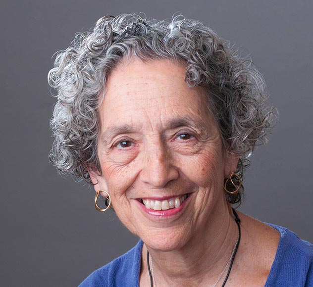 Ruth W. Messinger A smiling older woman with short, curly gray hair, wearing gold hoop earrings and a blue top. She is facing the camera against a gray background.