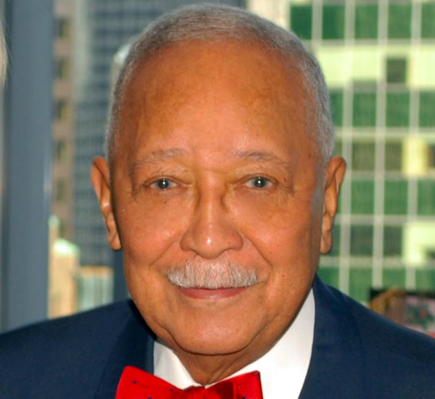 David Dinkins A man with short, gray hair and a mustache is wearing a blue suit and a red bow tie. He is smiling gently, and behind him are several high-rise buildings, slightly out of focus.