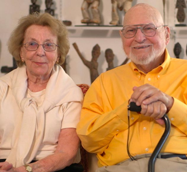 Fred and Rita Richman An elderly couple sits together smiling. The woman is wearing glasses and a light-colored blouse with a scarf draped over her shoulders. The man, also wearing glasses, has on a bright yellow shirt and holds a cane. A collection of sculptures is visible in the background.