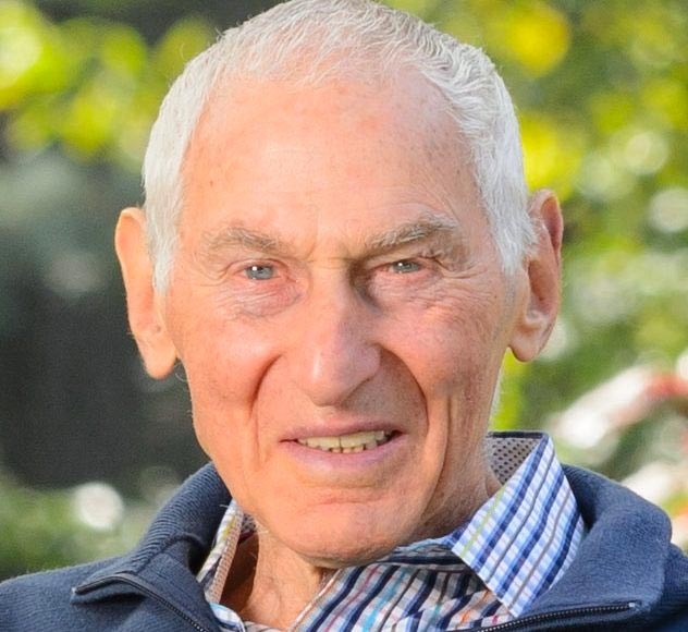 Harold Grinspoon An elderly man with short white hair smiles at the camera. He is wearing a blue zip-up jacket over a checkered shirt. The background is blurred, showing greenery and a hint of flowers.