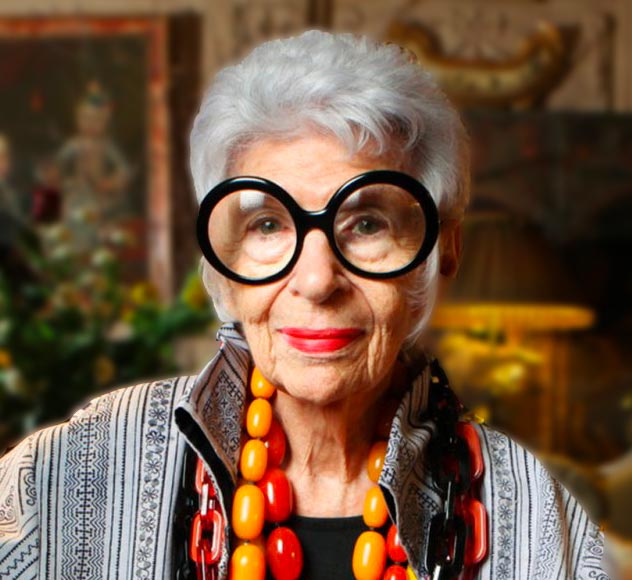 Iris Apfel An elderly woman with short white hair and large round black glasses is wearing a patterned jacket and vibrant chunky jewelry in orange and red. The background is softly blurred, featuring a lamp and some artwork or decorations.