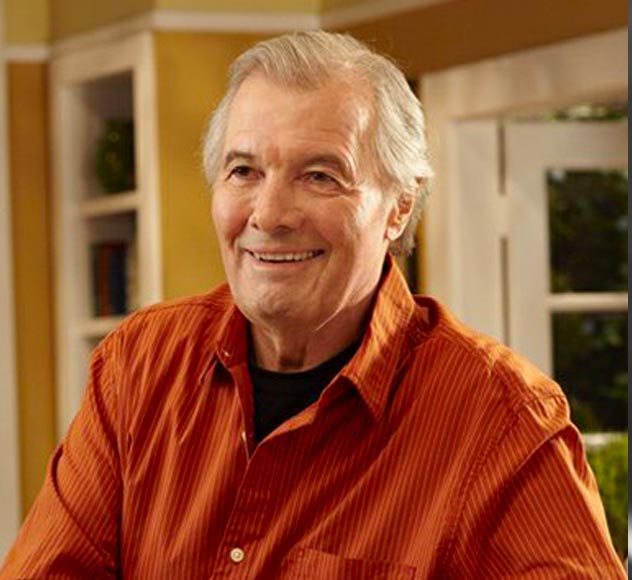 A smiling elderly man with gray hair wearing an orange shirt stands in a well-lit room with yellow and white walls, a window with white curtains, and a bookshelf in the background.