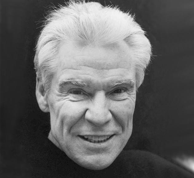 Jacques d’Amboise A black and white portrait of an elderly man with light-colored hair. He is smiling and wearing a dark, high-neck top. The background is dark, emphasizing his face.