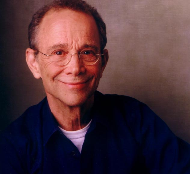 Joel Grey A smiling older man with short, light brown hair and glasses is wearing a dark blue shirt over a white undershirt. He is posed against a plain, neutral background.