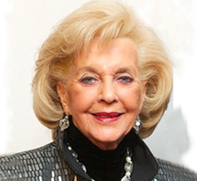 Joy Henshel An older woman with styled blonde hair is smiling at the camera. She is wearing a glittering jacket over a black turtleneck, accessorized with dangling earrings and a necklace. The background is light and simple.