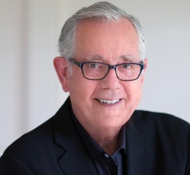Keith Reinhard An elderly man with short gray hair and glasses is smiling into the camera. He is wearing a charcoal blazer and a dark shirt. The background is plain and light-colored.