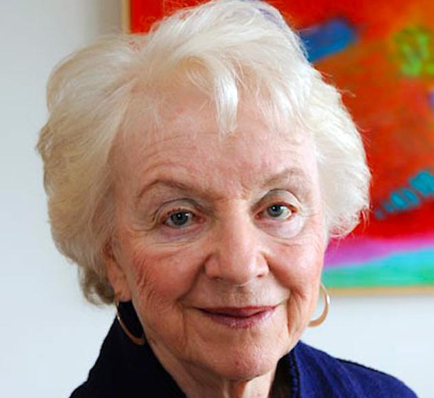 Madeleine May Kunin An elderly person with short, white hair and light skin smiles gently at the camera. They are wearing a dark-colored top and hoop earrings. The background features a colorful, abstract painting.