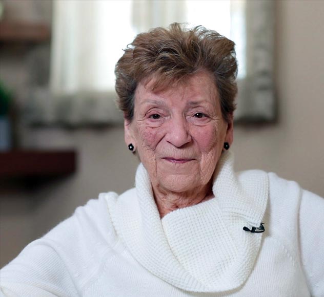 Marie Holley A smiling elderly woman with short, light brown hair is seated indoors. She is wearing a white sweater and small black earrings. Behind her, there is a soft-focus background with a window and a blurred shelf.