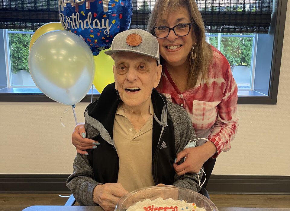 An elderly man and a woman smile together at a table with a birthday cake in front of them. The man is seated and the woman stands beside him with her arm around his shoulders. Blue and yellow balloons with "Happy Birthday" float above the cake.