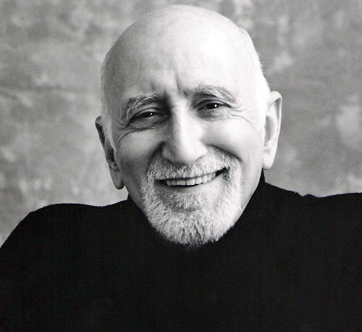 Dominic Chianese Black and white portrait of an elderly man with a shaved head and a white goatee. He is smiling warmly and wearing a dark turtleneck. The background is a textured, monochromatic backdrop.