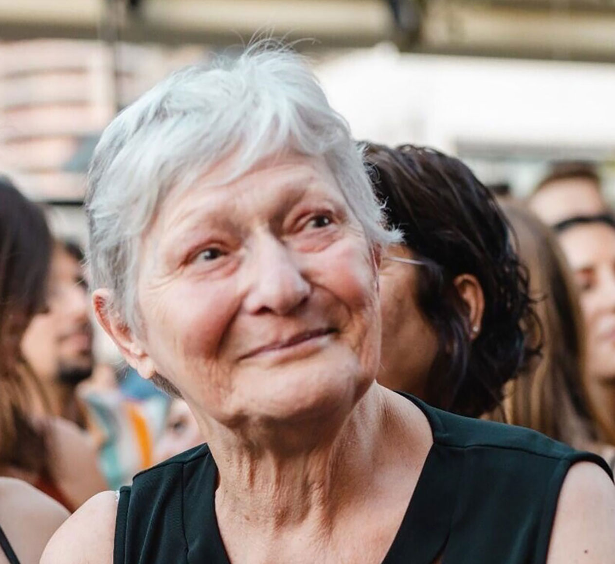 Yaffa-Adar- A smiling elderly woman with short white hair is wearing a sleeveless dark top. She is surrounded by a group of people in an outdoor setting, with blurred background elements suggesting a social gathering or event.