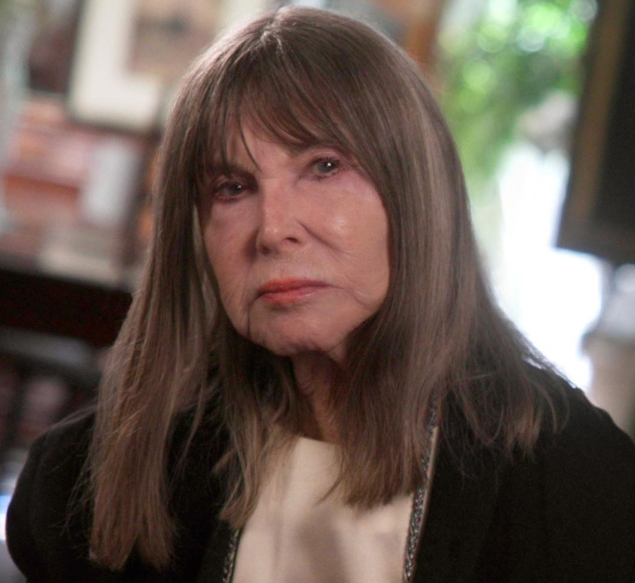 A woman with long, straight gray hair and bangs is looking slightly off-camera with a neutral expression. She is wearing a black jacket and a light-colored top. The background appears to be an indoor setting with blurred elements and greenery.