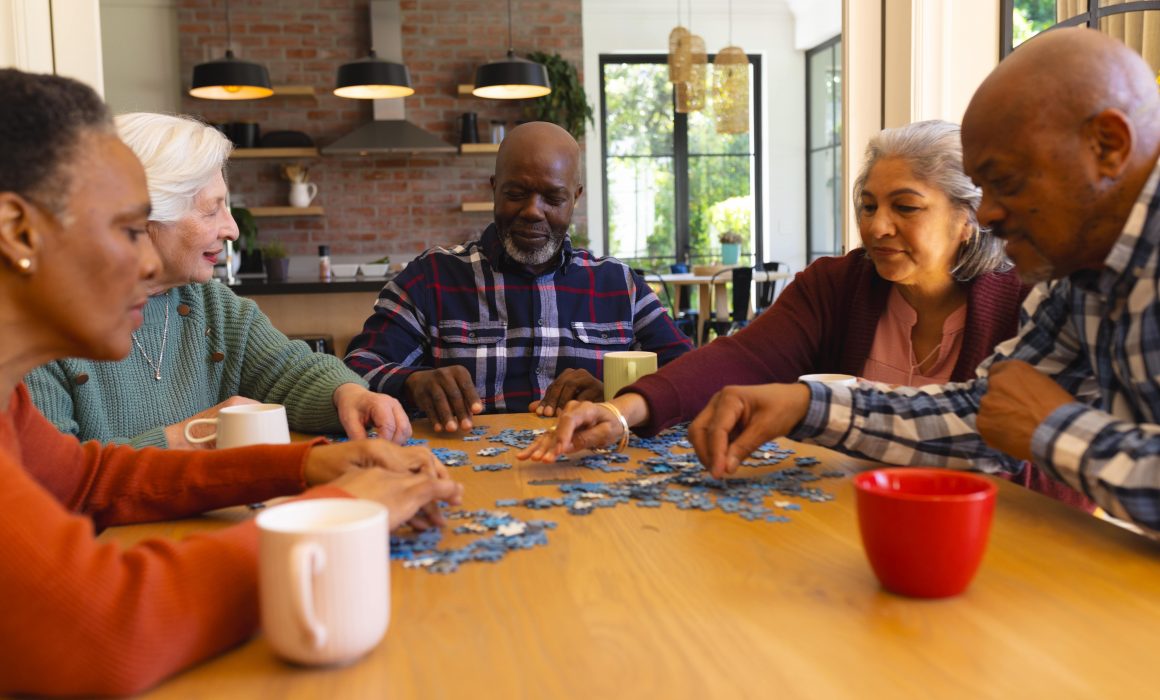 A group of five older adults is sitting around a wooden table in a cozy kitchen, working together on a jigsaw puzzle. They look focused and engaged. Coffee mugs are on the table, and a brick wall is visible in the background.