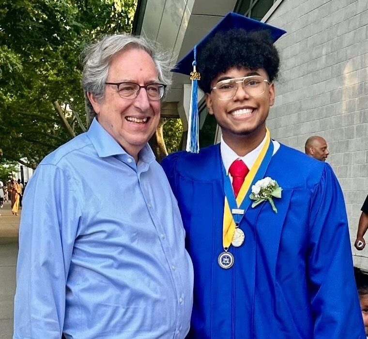 A young graduate in a blue cap and gown smiles while standing next to an older adult in a blue shirt and striped tie. They are outdoors, with trees and a building in the background. The graduate wears a medal and has a flower on their gown.