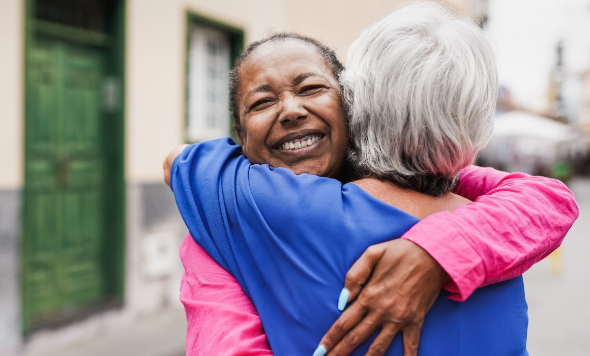 Two people embracing warmly on a street. The person facing the camera is smiling broadly and wearing a pink shirt, while the other, with gray hair, is facing away and wearing a blue shirt. A building with a green door is visible in the background.