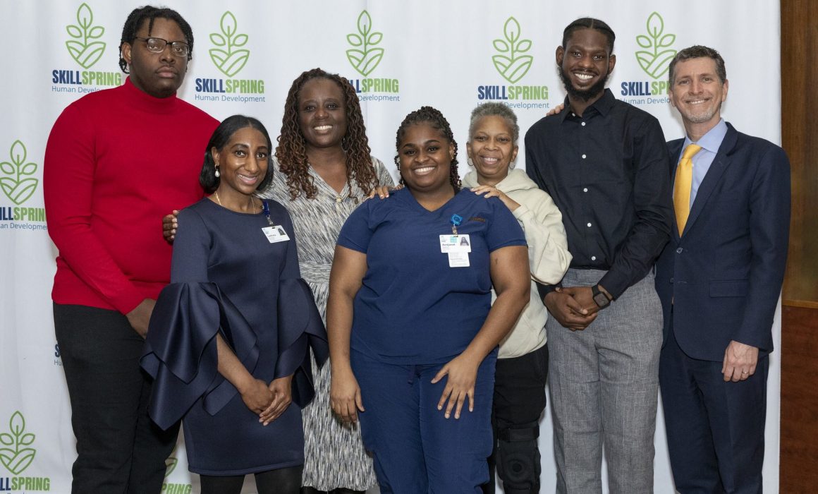 A group of seven people, six in business or casual attire and one in a blue medical uniform, stand smiling in front of a backdrop with the Skillspring Human Development logo featuring a green leaf design.