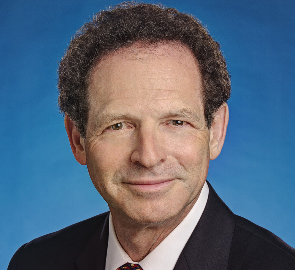 David-Freeman A person with curly brown hair wearing a dark suit, white shirt, and multicolored tie is smiling slightly against a blue background.