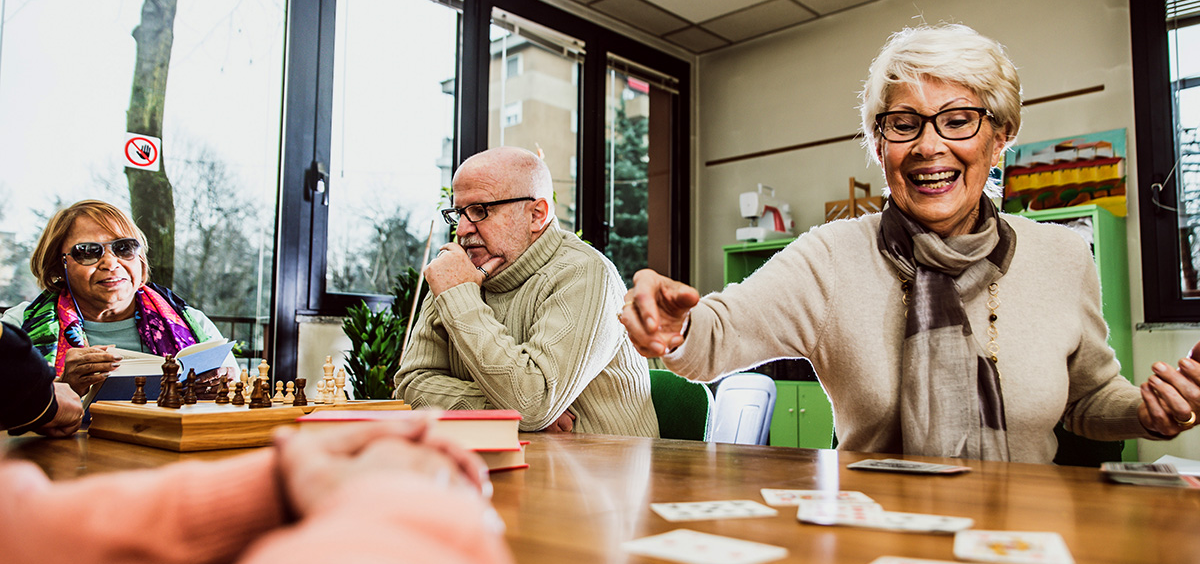 Hospitality-Program A group of older adults sits around a table playing cards and board games in a bright room; one woman is smiling and reaching for a card, while others chat and watch.