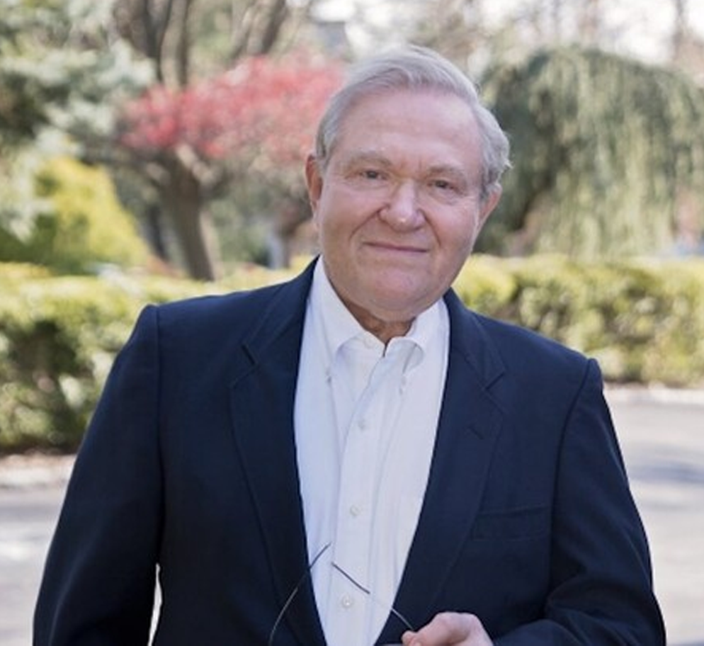 big-Michael Headshot Elderly man in a dark suit and white shirt stands outdoors holding glasses. Blurred trees and greenery in the background create a serene setting.