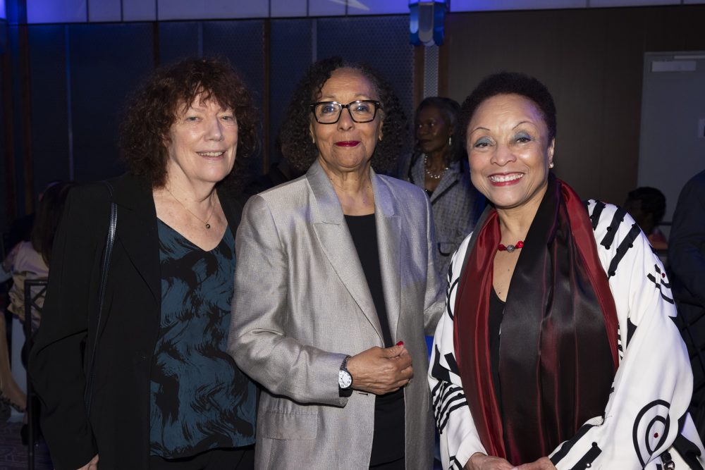 Three women posing together at an indoor event. The woman on the left has curly hair and wears a dark jacket, the middle woman wears glasses and a silver blazer, and the woman on the right wears a patterned jacket and smiles brightly.