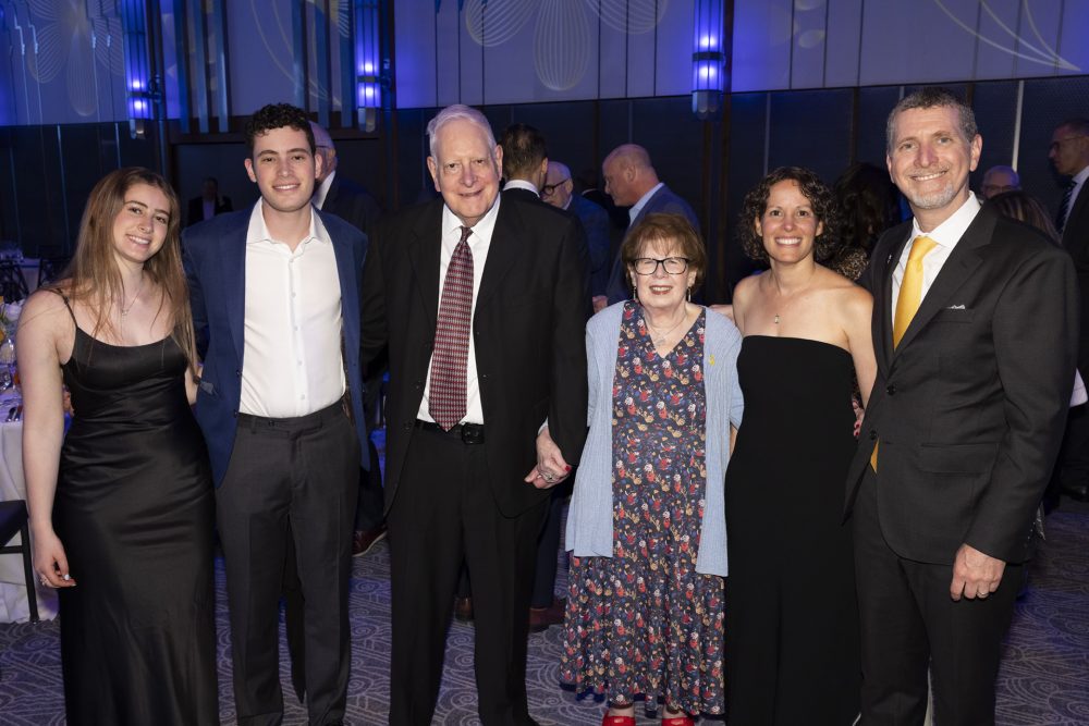 Six people dressed in formal attire stand together at an indoor event, smiling for the camera. The background is lit with blue lights and has other people mingling.