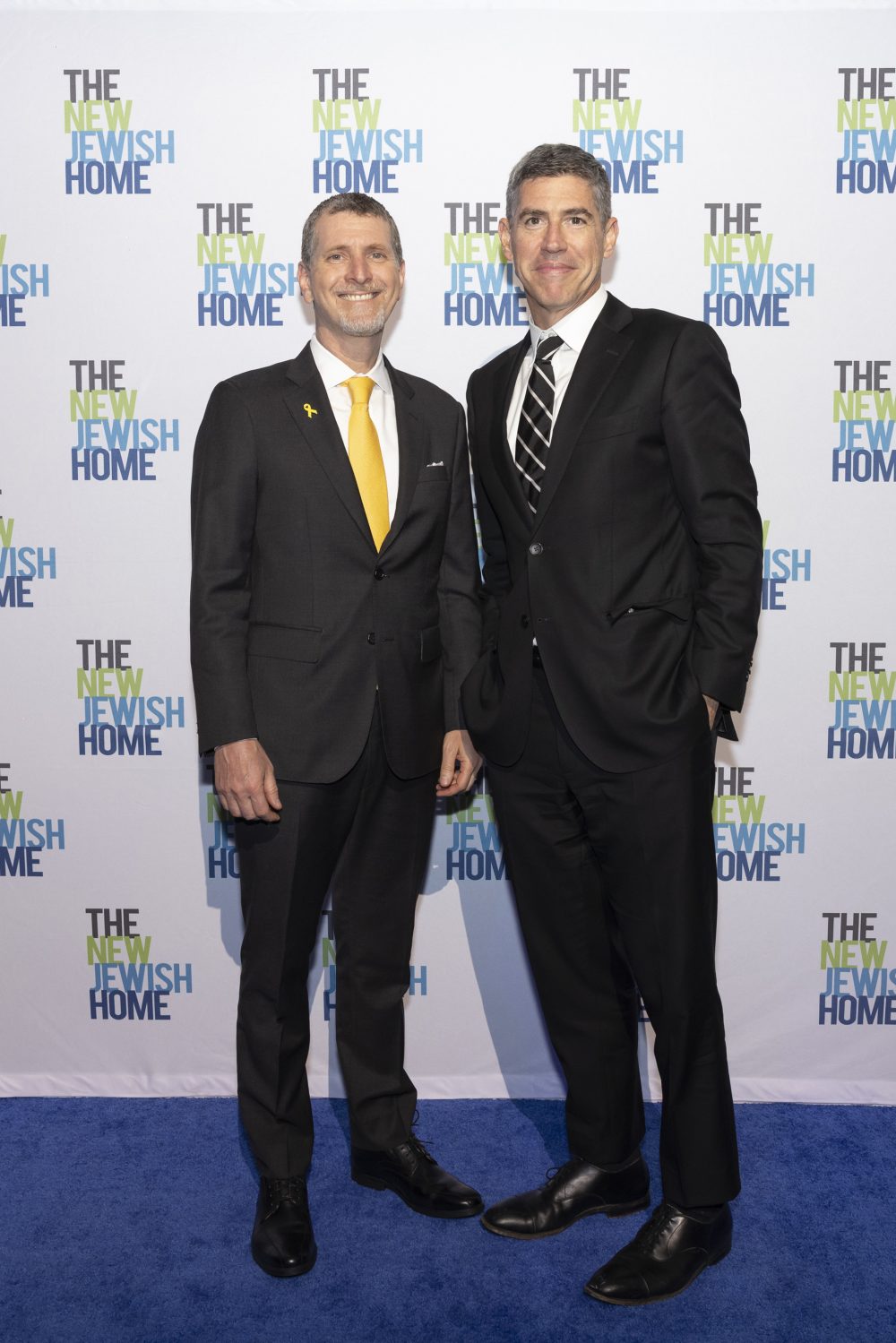 Two men in suits stand on a blue carpet in front of a backdrop that reads 