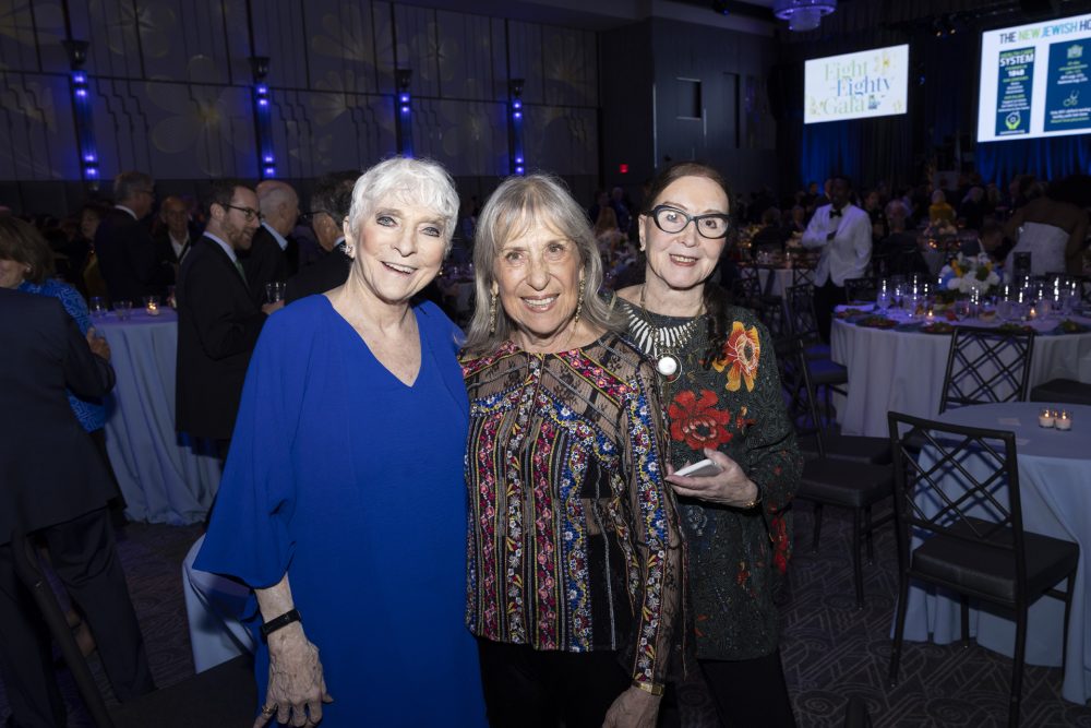 Three women pose and smile together at an indoor evening event, with decorated tables and other guests in the background under blue lighting. All three wear colorful, patterned outfits.