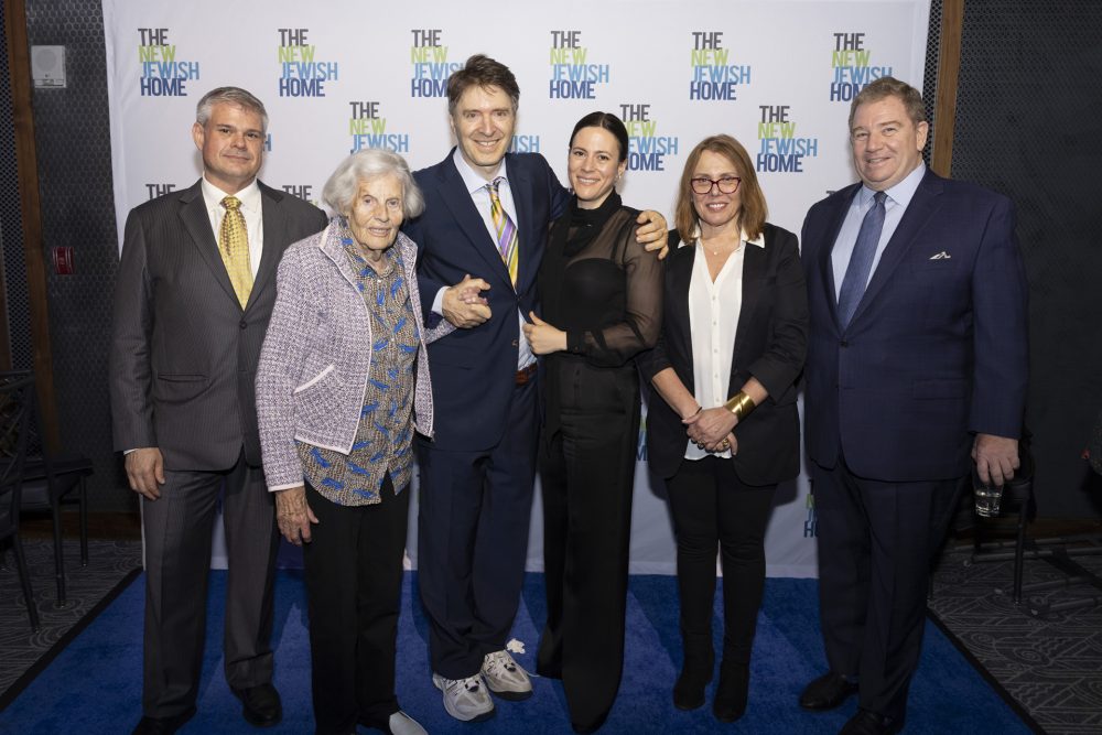 Six people stand together smiling in front of a step-and-repeat banner that says 