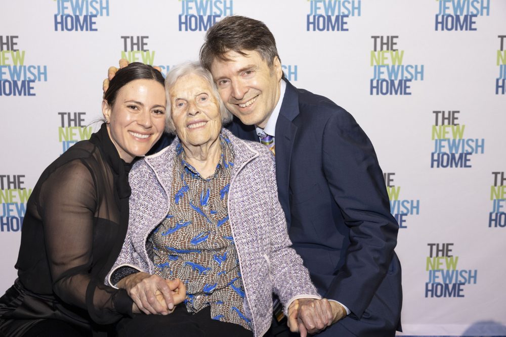 Three people smiling and posing together in front of a step-and-repeat banner that reads 