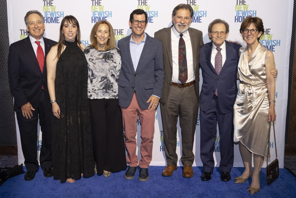 Seven people standing side by side, posing and smiling for a group photo on a blue carpet in front of a backdrop that reads 