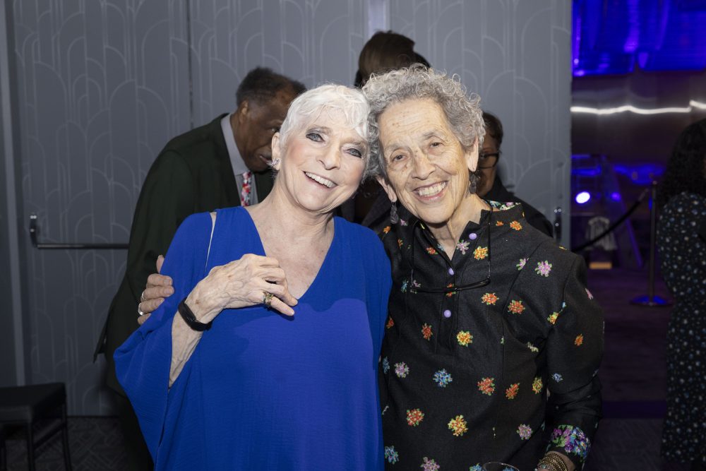 Two older women smiling and posing together at an indoor event, one wearing a blue dress and the other a black dress with colorful floral patterns, with people and patterned walls in the background.