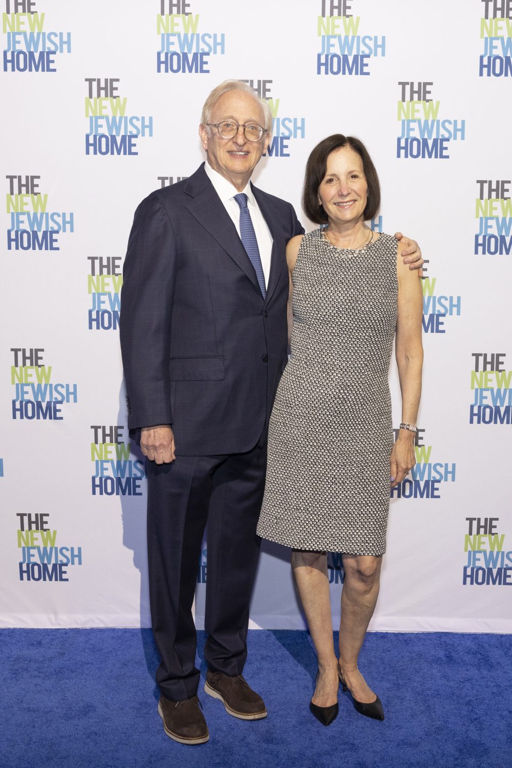 A man in a dark suit and tie stands with his arm around a woman in a sleeveless patterned dress. They both smile at the camera on a blue carpet in front of a backdrop that says 