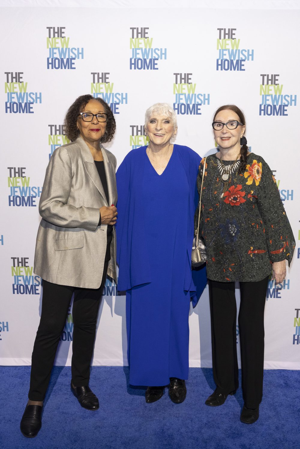 Three women stand together on a blue carpet in front of a step-and-repeat backdrop with 