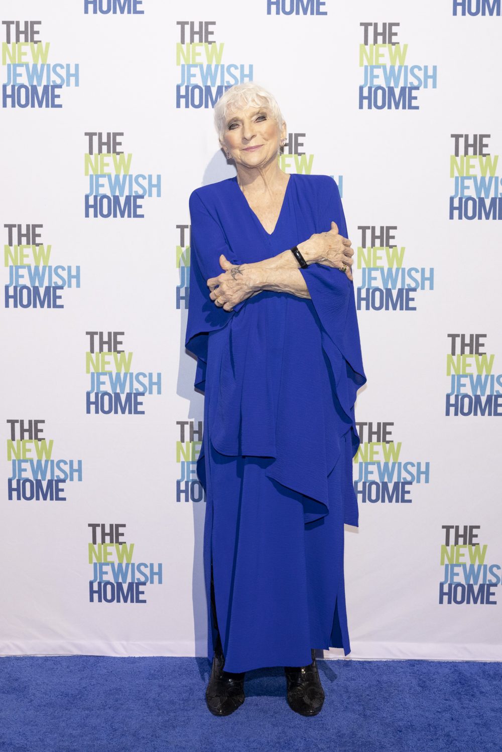 An older woman in a long, flowing blue dress stands on a blue carpet, crossing her arms and smiling. She poses in front of a step-and-repeat backdrop with “THE NEW JEWISH HOME” repeated in various colors.