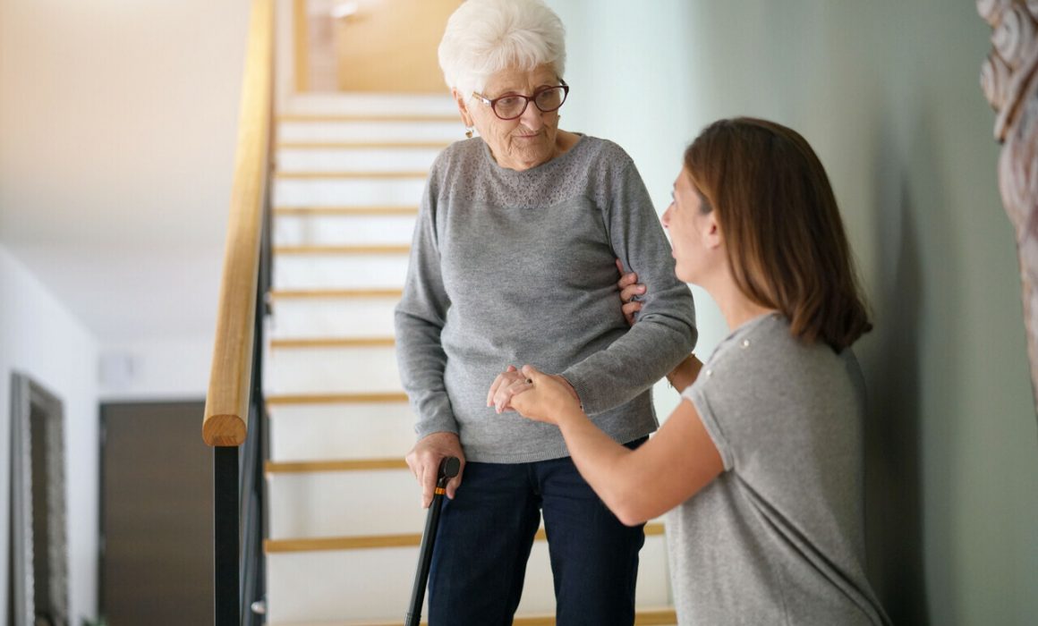 A young woman assists an elderly woman with glasses and a cane as she walks down a staircase in a home, holding her arm for support.