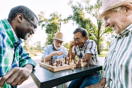 Four older men happily play chess together outdoors at a table, laughing and enjoying each other's company on a sunny day, with trees and greenery in the background.